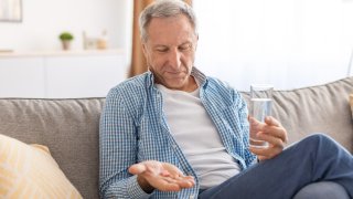 Foto ilustrativa de un hombre mayor sentado en un sofá con un vaso de agua, sosteniendo pastillas en la mano tomando analgésicos en casa.