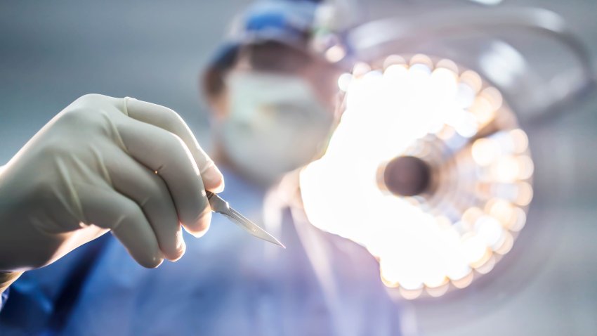 Doctor or surgeon in blue uniform holding surgical knife or scalpel to do surgery inside operating room in hospital under surgical lamp.People pick up surgical blade with white clean space.